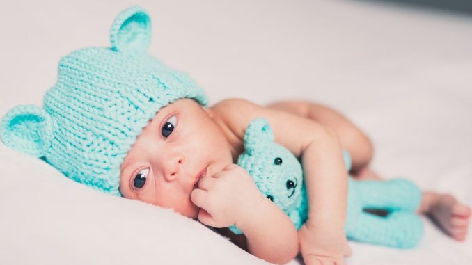 Un nourrisson avec bonnet d'ours et peluche bleue sur drap blanc